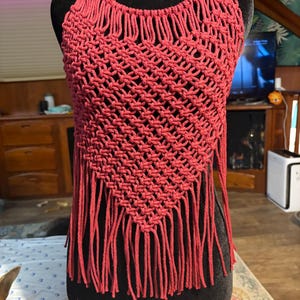 May include: A handmade, red macrame halter top with a diamond-shaped pattern and fringe detail. The top is displayed on a black mannequin. The halter top is a unique piece of bohemian-inspired clothing.