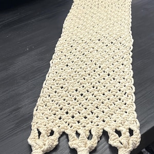 May include: Cream-colored macrame table runner with a textured, open weave design. The runner features decorative knotted tassels at each end and a scalloped edge. This handmade textile adds a bohemian touch to home decor.