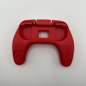May include: A bright red 3D-printed controller grip stand featuring a recessed central area for device placement and a small leaf-shaped icon embossed on the left handle. The surface shows horizontal layer lines from the printing process.