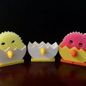 May include: Three decorative chick-shaped sponge holders. Each holder is a white eggshell with a yellow or orange base and beak. The sponges are yellow, white, and pink, with eye cutouts, resembling chicks.