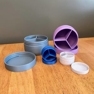May include: A set of six small, round containers with lids. The containers are made of plastic and have three compartments each. The containers are in various colors, including purple, blue, and gray.