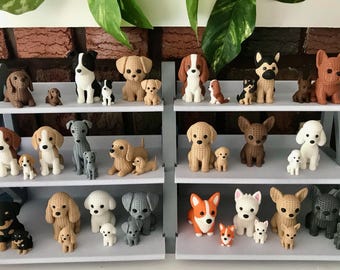 Könnte beinhalten: Eine Sammlung kleiner, gehäkelter Hundefiguren, die auf einem dreistufigen grauen Regal ausgestellt sind. Das Set umfasst verschiedene Rassen in unterschiedlichen Farben wie Braun, Schwarz, Weiß, Grau und Hellbraun, wobei jeder große Hund von einem Welpen begleitet wird.