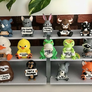 May include: A collection of thirteen small animal figurines arranged on a three-tiered gray display stand. Each figure holds a white sign with humorous text, including phrases like 'No treats No Kisses', 'Warning I Thump', 'Duck Off', and 'Not a Sausage'.