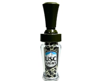 May include: A green and black camouflage duck call with a clear plastic body and a green plastic top. The call has the text "USC LIGHT" printed on the side.