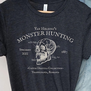 May include: Dark grey heather t-shirt with the text "Van Helsing's Monster Hunting" and a detailed skull graphic. The shirt also includes the text "Specimen XIII" and "1887". The bottom of the shirt reads "Castle Dracula Collection, Transylvania, Romania".