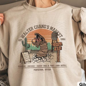 Puede incluir: Sudadera de color arena con un gráfico que presenta una escena desértica con el texto "WALTER CHANG'S MARKET" y "PERFECTION, NEVADA". El diseño incluye un cactus, una torre de agua y una criatura. La sudadera también tiene el texto "THE GEEK CHIC CO."