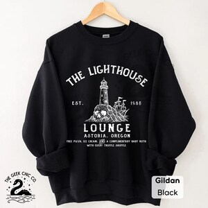 May include: Black sweatshirt with a white graphic featuring a lighthouse, skull, and ship, with the text "The Lighthouse Lounge Astoria, Oregon." The sweatshirt also has the text "Est. 1985" and "Free Pizza, Ice Cream, and a Complimentary Baby Ruth with Every Truffle Shuffle."