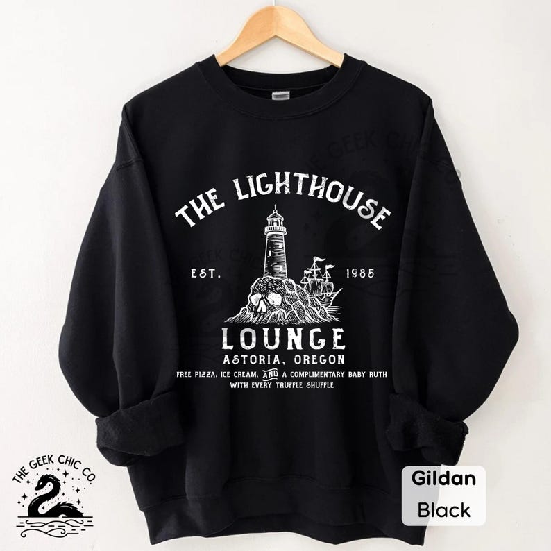 May include: Black sweatshirt with a white graphic featuring a lighthouse, skull, and ship, with the text "The Lighthouse Lounge Astoria, Oregon." The sweatshirt also has the text "Est. 1985" and "Free Pizza, Ice Cream, and a Complimentary Baby Ruth with Every Truffle Shuffle."