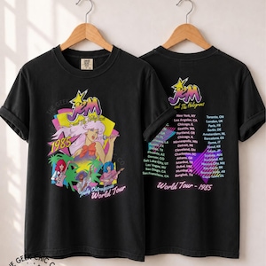 May include: A black t-shirt featuring a graphic of Jem and the Holograms with the text 1985 and Truly Outrageous World Tour on the front, and a list of global tour cities with the text World Tour - 1985 on the back, both hanging on wooden hangers.