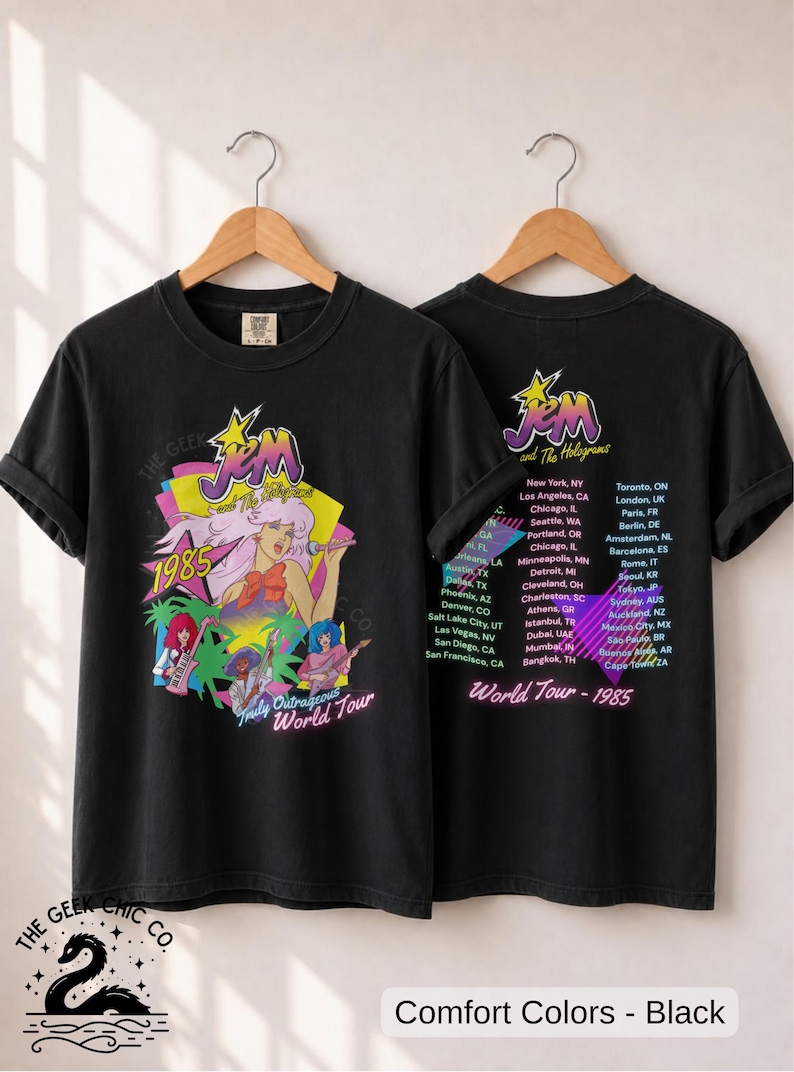 May include: A black t-shirt featuring a graphic of Jem and the Holograms with the text 1985 and Truly Outrageous World Tour on the front, and a list of global tour cities with the text World Tour - 1985 on the back, both hanging on wooden hangers.