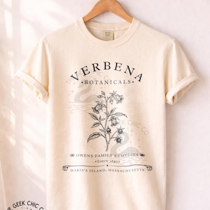 Könnte beinhalten: Ein elfenbeinfarbenes Kurzarm-T-Shirt mit einer schwarzen botanischen Illustration einer blühenden Pflanze. Der Text lautet VERBENA BOTANICALS, OWENS FAMILY REMEDIES, Since 1680 und MARIA'S ISLAND, MASSACHUSETTS. Das T-Shirt hängt an einem Holzkleiderbügel.