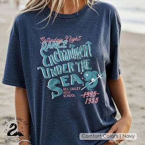 May include: Navy blue t-shirt with the text "Saturday Night Dance Enchantment Under The Sea" in a retro font. Features a dolphin graphic and the years 1985 and 1955. The shirt is from Hill Valley High School.