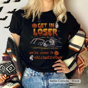 Pode incluir: Camiseta preta com um design de Halloween. O design apresenta o texto "GET IN LOSER" acima de um carro dirigido por um esqueleto, com a frase "WE'RE GOING TO HALLOWEENTOWN" abaixo. Morcegos, abóboras e um fantasma também estão incluídos.