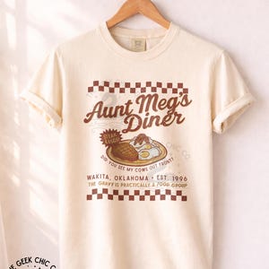 May include: An ivory short-sleeve t-shirt featuring a brown graphic of a diner meal with steak and eggs. The text reads Aunt Meg's Diner, Steak & Eggs, Did you see my cows out front?, Wakita, Oklahoma, Est. 1996, and The gravy is practically a food group.