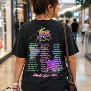May include: A black short-sleeve t-shirt featuring a graphic print on the back with the text Jem and The Holograms, World Tour 1985, and a list of global cities. The design includes a yellow star, purple lettering, and pink geometric shapes.