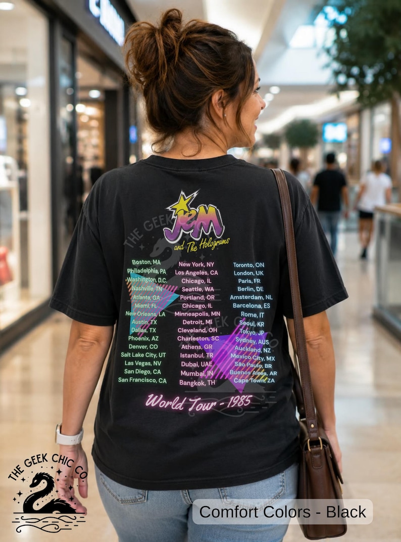 May include: A black short-sleeve t-shirt featuring a graphic print on the back with the text Jem and The Holograms, World Tour 1985, and a list of global cities. The design includes a yellow star, purple lettering, and pink geometric shapes.