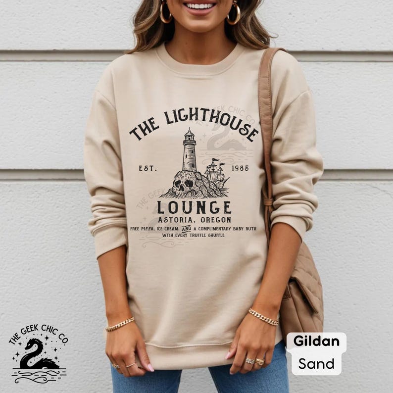 May include: Sand-colored sweatshirt with a graphic that reads "The Lighthouse Lounge Astoria, Oregon" featuring a lighthouse, skull, and ship design. The sweatshirt also has the text "Est. 1985" and "Free Pizza, Ice Cream, and a Complimentary Baby Ruth with Every Truffle Shuffle."