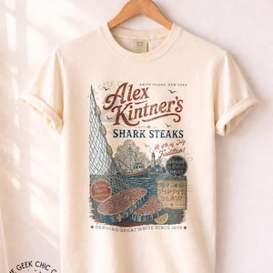 May include: An ivory short-sleeve t-shirt featuring a graphic print with the text Alex Kintner's Shark Steaks, Amity Island, New York, A 4th of July Tradition, Fresh Cut Daily, and Serving Great White Since 1975, alongside illustrations of a fishing boat, net, and steaks.