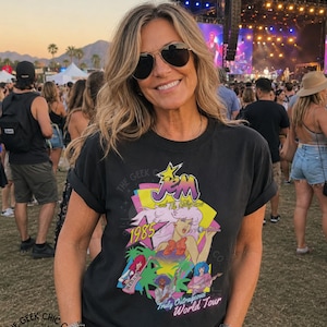 May include: A black short-sleeve t-shirt featuring a colorful graphic print of Jem and the Holograms with the text 1985 and Truly Outrageous World Tour. The design includes stylized characters, a yellow star, and palm trees on a dark background.