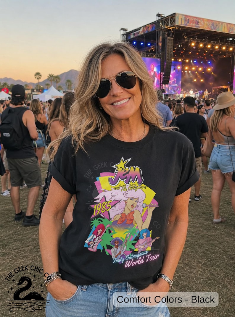 May include: A black short-sleeve t-shirt featuring a colorful graphic print of Jem and the Holograms with the text 1985 and Truly Outrageous World Tour. The design includes stylized characters, a yellow star, and palm trees on a dark background.
