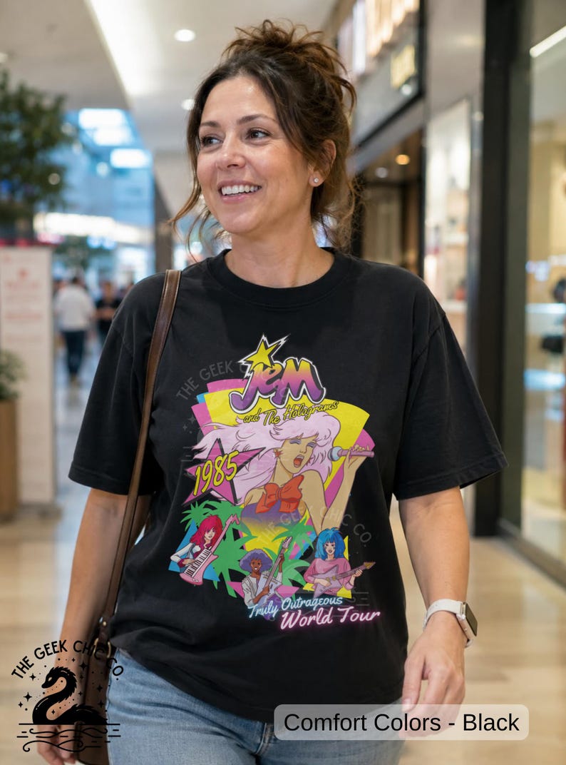 May include: A black short-sleeve t-shirt featuring a colorful graphic of Jem and the Holograms with the text Jem and the Holograms, 1985, and Truly Outrageous World Tour. The design includes stylized illustrations of band members and musical instruments.