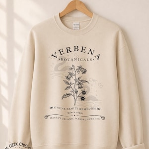 May include: A sand-colored crewneck sweatshirt featuring a black botanical illustration of a flowering plant. The text reads VERBENA BOTANICALS, OWENS FAMILY REMEDIES, Since 1680, and MARIA'S ISLAND, MASSACHUSETTS. A faint swan graphic is visible in the background.