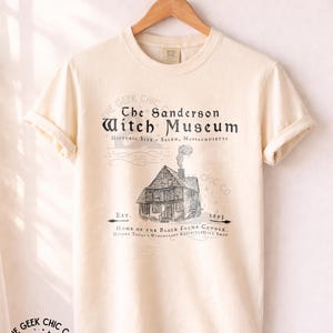 May include: An ivory short-sleeve t-shirt featuring a black graphic print of a house with smoke rising from the chimney. The text reads: The Sanderson Witch Museum, Historic Site - Salem, Massachusetts, Est. 1693, Home of the Black Flame Candle.