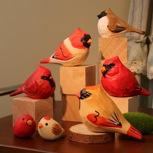 以下が含まれることがあります： 手彫りの木製鳥の置物のコレクション。鳥は赤、茶色、黄色などの鮮やかな色で塗装されており、黒のアクセントが付いています。一部の鳥は小さな木のブロックに止まり、他の鳥は木の輪切りに止まっています。コレクションには、カージナルスやその他の鳥種が含まれています。