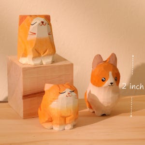 May include: Three wooden cat figurines, one on a wooden block, and a corgi figurine. The cat figurines are orange and white, and the corgi is orange and white. The corgi is approximately 2 inches tall.