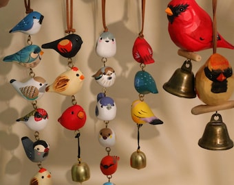 May include: A collection of colorful, hand-painted wooden bird ornaments. Each bird is unique, with varying colors and designs, and hangs from a leather cord. Some have small brass bells attached. The birds include blue, red, yellow, and white varieties.