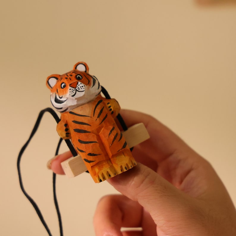 K&ouml;nnte beinhalten: Handgeschnitzte Holz-Tigerfigur mit orangefarbenen und schwarzen Streifen, wei&szlig;en Akzenten und einem l&auml;chelnden Gesicht. Der Tiger ist an einer kleinen Holzschaukel mit schwarzen Schn&uuml;ren befestigt. Die Figur wird von einer Person gehalten.