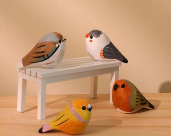 Puede incluir: Cuatro figuras de pájaros de madera pintadas a mano. Uno es amarillo con detalles rosas, otro es naranja y gris, y dos están posados en un pequeño banco blanco. Los pájaros tienen detalles y están sobre una superficie de madera.