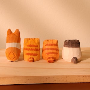 May include: Four hand-carved wooden cat figurines. The figurines are orange, white, and brown, with details like stripes and ears. They are displayed on a light-colored wooden surface, showcasing the craftsmanship.