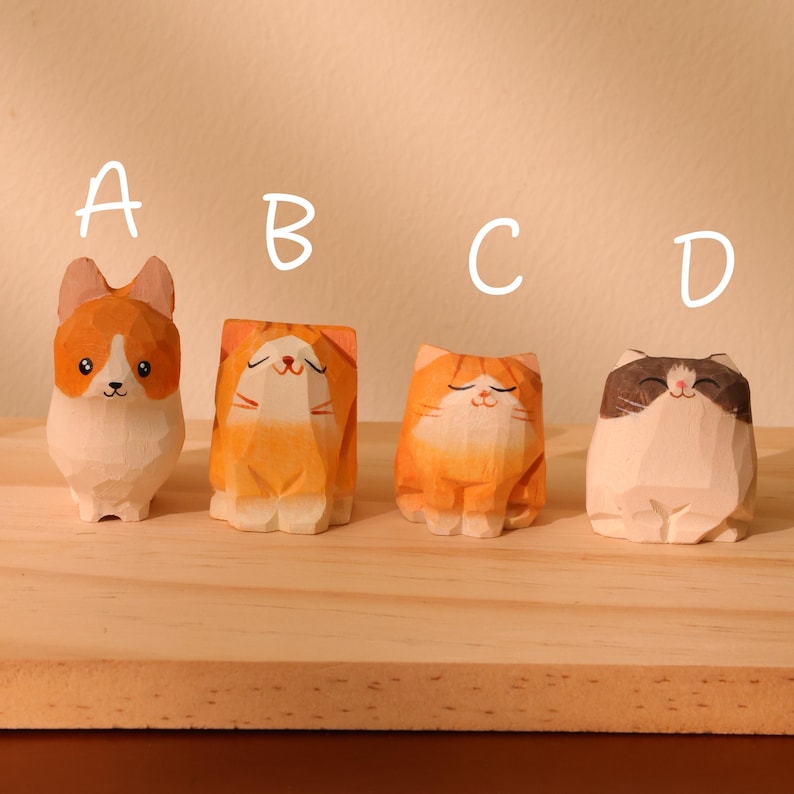 May include: Four hand-carved wooden figurines of animals, including a corgi and three cats. The corgi is white and orange, while the cats are orange, and white and brown. Each figurine has a unique, smiling expression and is displayed on a wooden surface.