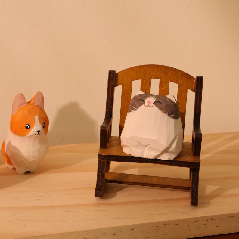 May include: Wooden carved figurines of a corgi and a cat. The cat is seated in a miniature wooden chair. The corgi is orange and white, and the cat is white and brown. The figurines are on a light-colored wooden surface.