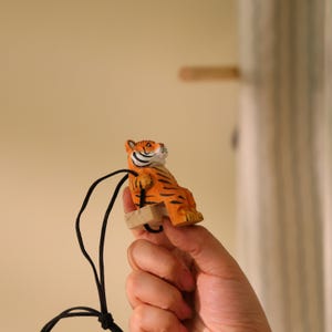 K&ouml;nnte beinhalten: Eine kleine, orange und schwarz gestreifte Tigerfigur mit schwarzem Band. Der Tiger sitzt auf einer kleinen Holzschaukel. Die Figur wird von einer Hand gehalten.