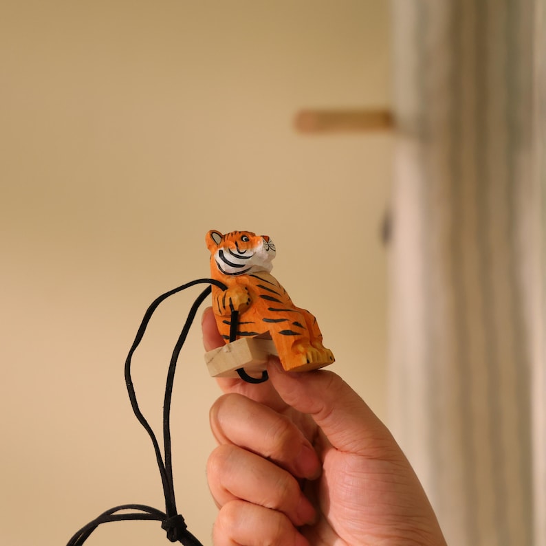 K&ouml;nnte beinhalten: Eine kleine, orange und schwarz gestreifte Tigerfigur mit schwarzem Band. Der Tiger sitzt auf einer kleinen Holzschaukel. Die Figur wird von einer Hand gehalten.