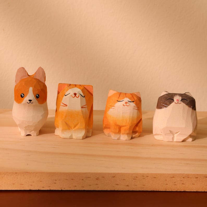 May include: Four wooden cat figurines in various poses and colors. The figurines include a corgi and three cats, each with unique expressions and markings. The corgi is white and orange, while the cats are orange, and white and brown. They are displayed on a wooden surface.