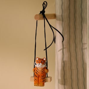 K&ouml;nnte beinhalten: Eine Holzfigur eines Tigers auf einer Schaukel. Der Tiger ist orange mit schwarzen Streifen und einem wei&szlig;en Gesicht. Die Schaukel h&auml;ngt an schwarzen Schn&uuml;ren von einem Holzd&uuml;bel. Der Hintergrund ist eine hellbeige Wand und ein gestreifter Vorhang.