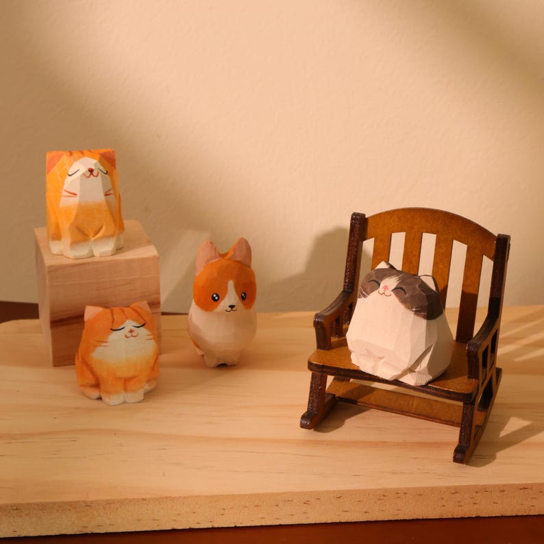 May include: Wooden cat figurines in orange, white, and brown. One cat sits in a miniature wooden rocking chair. Two cats are on a wooden block, and another stands on a wooden surface. The figurines have a carved, geometric design.