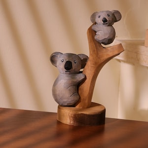 Può includere: Scultura in legno di due koala su un ramo d'albero. I koala sono grigi con nasi e occhi neri. Il ramo e la base sono marroni. La scultura è intagliata a mano e ha un aspetto rustico.