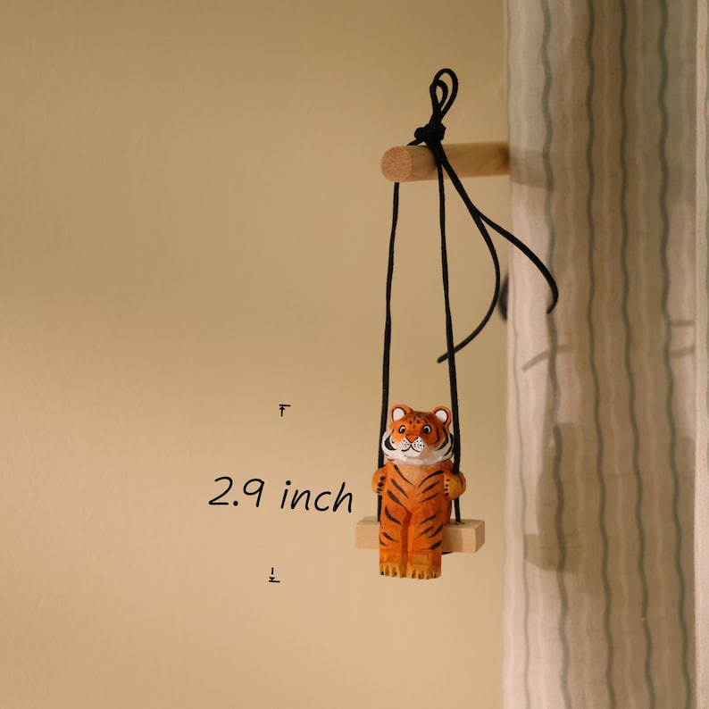 K&ouml;nnte beinhalten: Eine Holz-Tigerfigur auf einer Schaukel. Der Tiger ist orange mit schwarzen Streifen und einem wei&szlig;en Gesicht. Die Schaukel ist mit schwarzen Schn&uuml;ren an einem Holzd&uuml;bel aufgeh&auml;ngt. Der Tiger ist 7,37 cm gro&szlig;.