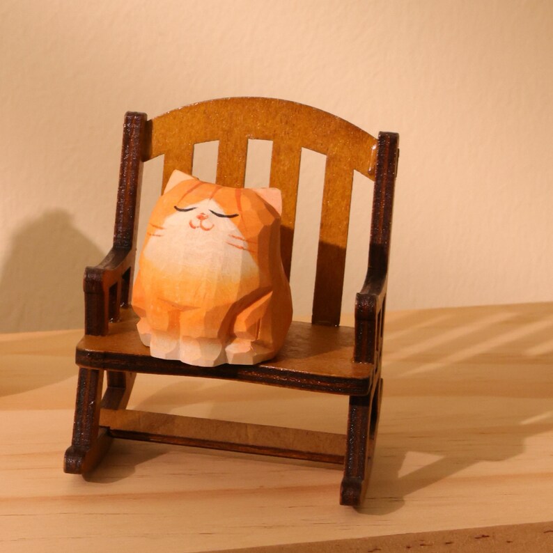 May include: A wooden carving of a relaxed orange and white cat sitting in a miniature wooden rocking chair. The cat has closed eyes and a peaceful expression. The chair is brown with a slatted back.