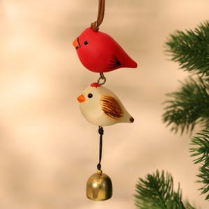 May include: A hanging ornament featuring two bird figurines, one red and one cream-colored, connected by a metal ring. The cream-colored bird has brown wing markings and a small brass bell hangs below. The ornament is suspended by a brown leather cord.