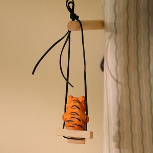 K&ouml;nnte beinhalten: Eine Holzfigur eines Tigers mit orangefarbenen und schwarzen Streifen sitzt auf einer kleinen Holzschaukel, die an schwarzen Schn&uuml;ren h&auml;ngt. Die Schaukel ist an einem Holzd&uuml;bel befestigt. Der Hintergrund ist ein neutrales Beige.