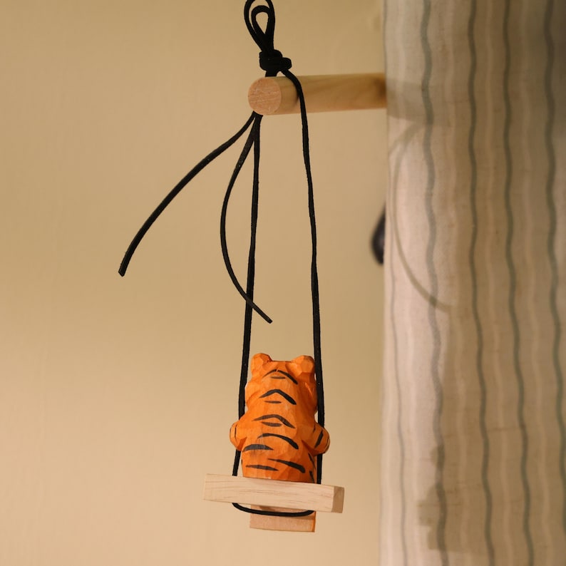 K&ouml;nnte beinhalten: Eine Holzfigur eines Tigers mit orangefarbenen und schwarzen Streifen sitzt auf einer kleinen Holzschaukel, die an schwarzen Schn&uuml;ren h&auml;ngt. Die Schaukel ist an einem Holzd&uuml;bel befestigt. Der Hintergrund ist ein neutrales Beige.