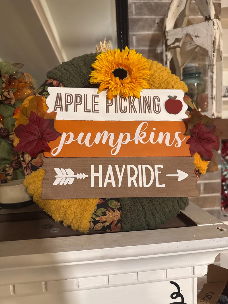 以下が含まれることがあります： 「Apple Picking pumpkins hayride」と書かれた木製の看板が付いた秋のリース。リースは緑、茶色、黄色の布で作られており、上にひまわりが飾られています。