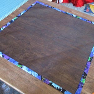 May include: A dark brown wooden board with a floral patterned border in shades of blue, purple, and green. The board is rectangular and appears to be sitting on a wooden surface.