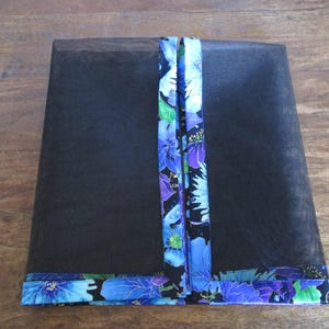 May include: Black mesh cover with a floral patterned trim and accents. The trim and vertical accents feature blue, purple, and green flowers on a black background. The cover is rectangular and appears to be for a food item.