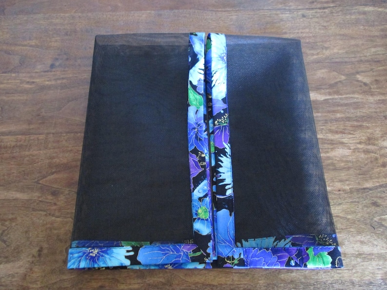 May include: Black mesh cover with a floral patterned trim and accents. The trim and vertical accents feature blue, purple, and green flowers on a black background. The cover is rectangular and appears to be for a food item.